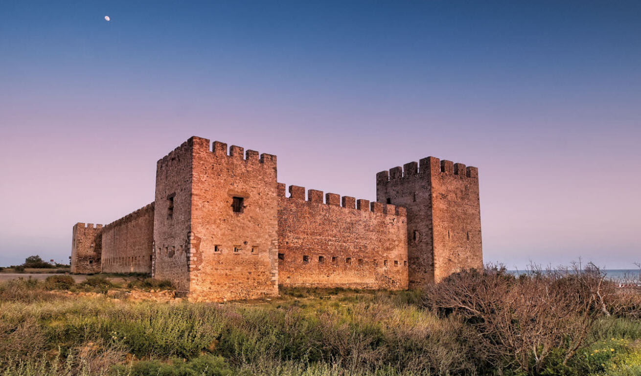 Large stone castle with turrets and loopholes at sunset. Around low vegetation and in the background the sea.