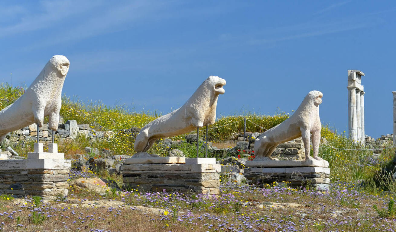 3 ancient lion statues in a row and in the background 4 ancient columns with low vegetation and wild flowers.