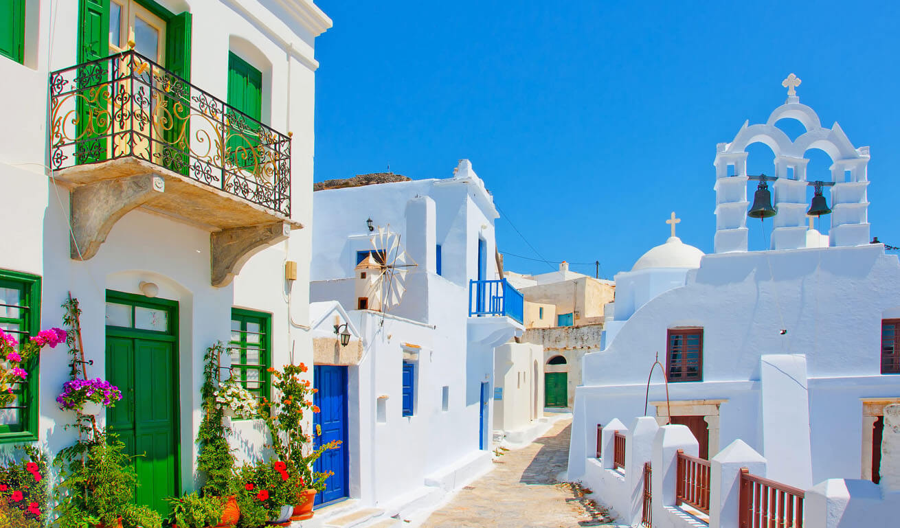 Κυκλαδίτικο καλντερίμι με λευκά σπίτια και εκκλησία. Πολλές γλάστρες με λουλούδια στην είσοδο του ενός σπιτιού.