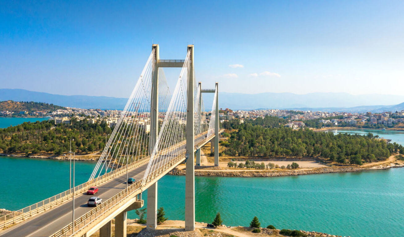 Modern cable bridge over the sea that leads to a tree-lined area. In the background you can see a city.