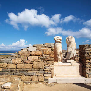 Archaeological site with low stone walls and 2 headless statues. In the background is the sea.