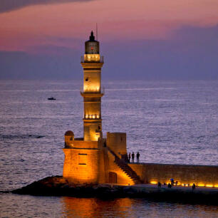 Old lighted lighthouse made of stone on the edge of a pier with a few people at sunset.