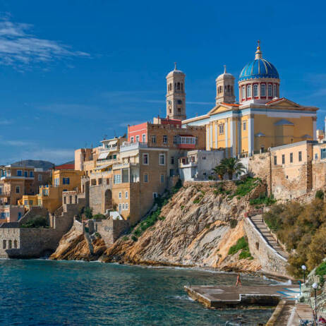 Μεγάλη χρωματιστή εκκλησία με μπλε τρούλο πάνω από χρωματιστά σπίτια που φτάνουν ως τη θάλασσα. Μικρή προβλήτα μπροστά.