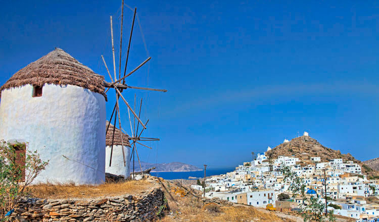 2 windmills at the top of a hill and in the background a traditional Cycladic settlement
