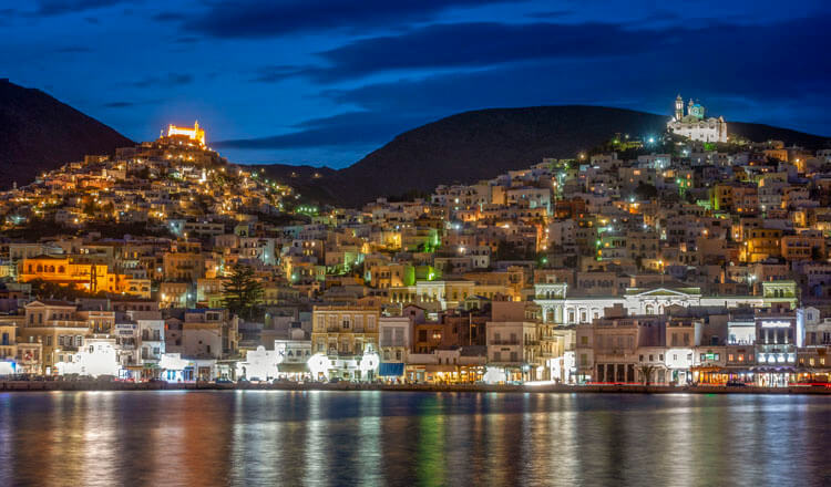 Night view of a large settlement from the sea with hills that have churches on the tops.