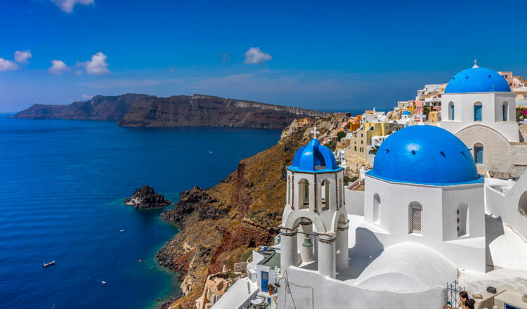 The domes of 2 churches and a settlement on a high rock above the sea and in the background 2 small islands nearby.