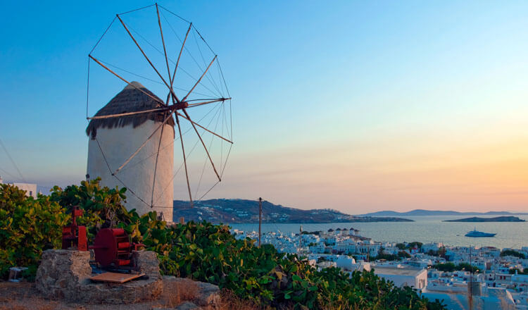 Windmill next to low vegetation above a traditional settlement with white houses and in the background the sea at sunset.