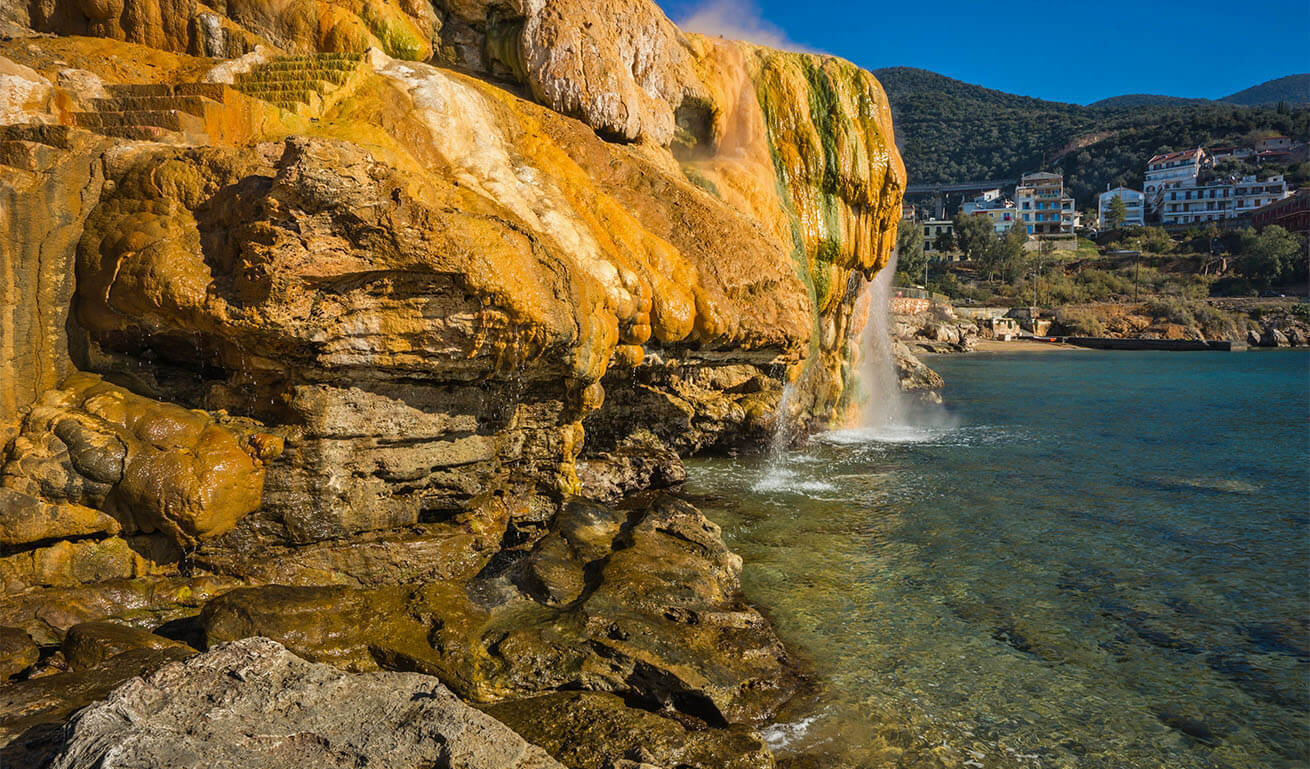 Seaside rocks with a yellowish tint and water forming a small waterfall. In the background, a green slope with a few houses that ends at a beach. Sunny day.
