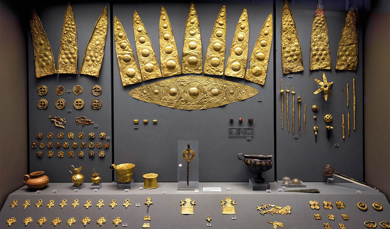 Museum display case with embossed gold objects and jewelry of various sizes.