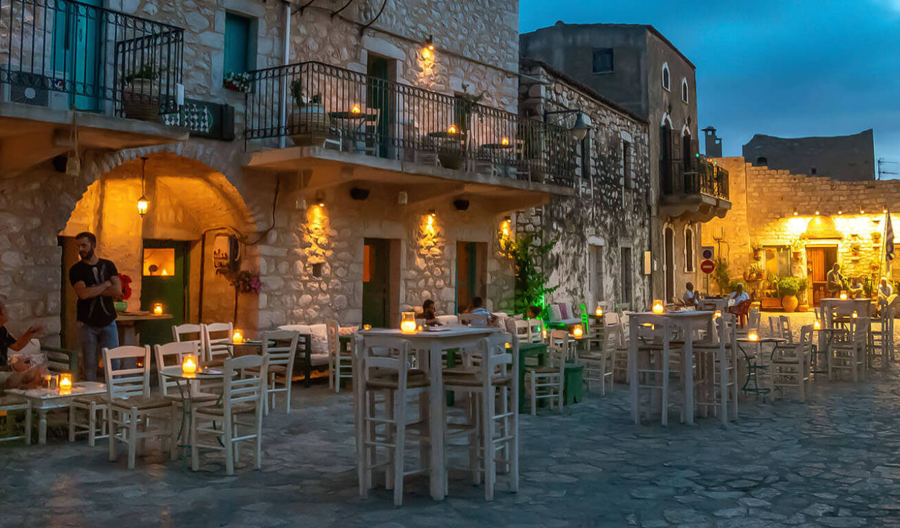 Cobblestone square with wooden tables and chairs in front of stone two- and three-story buildings at dusk with lights on.