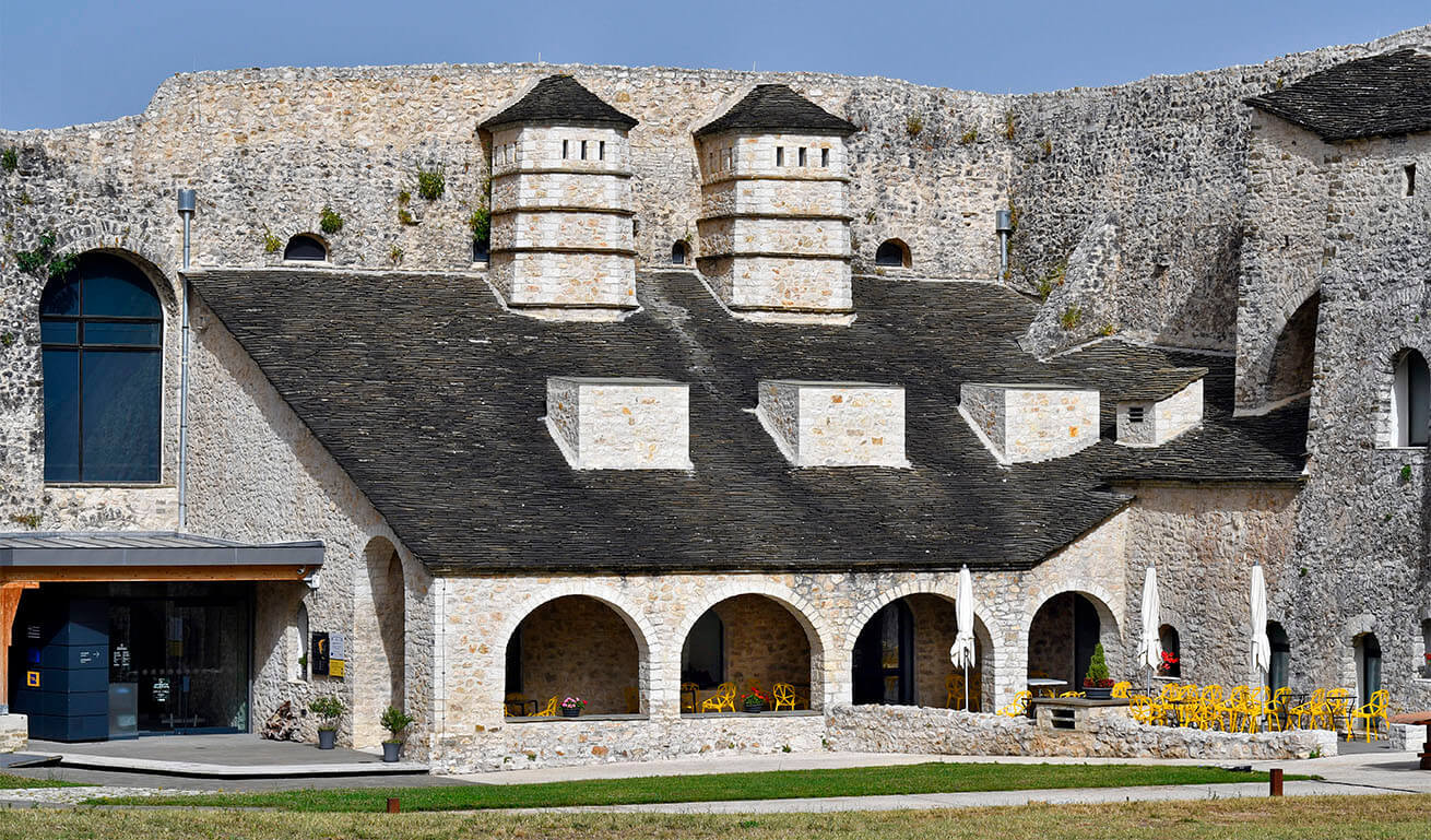 Stone building with arched windows, slate roof, twin chimneys, and an outdoor seating area with yellow chairs and white umbrellas.