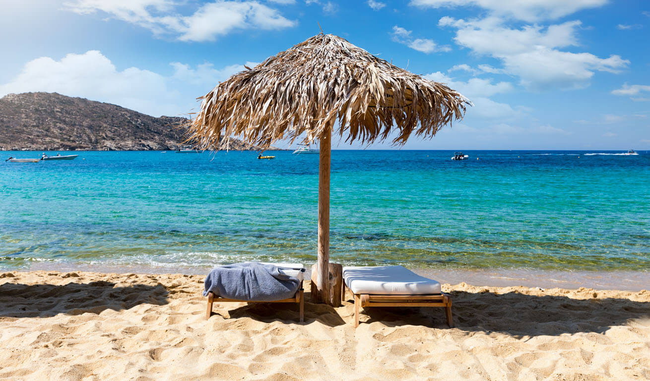 2 comfortable sunbeds in the shade of an umbrella on a sandy beach in front of the calm sea. 