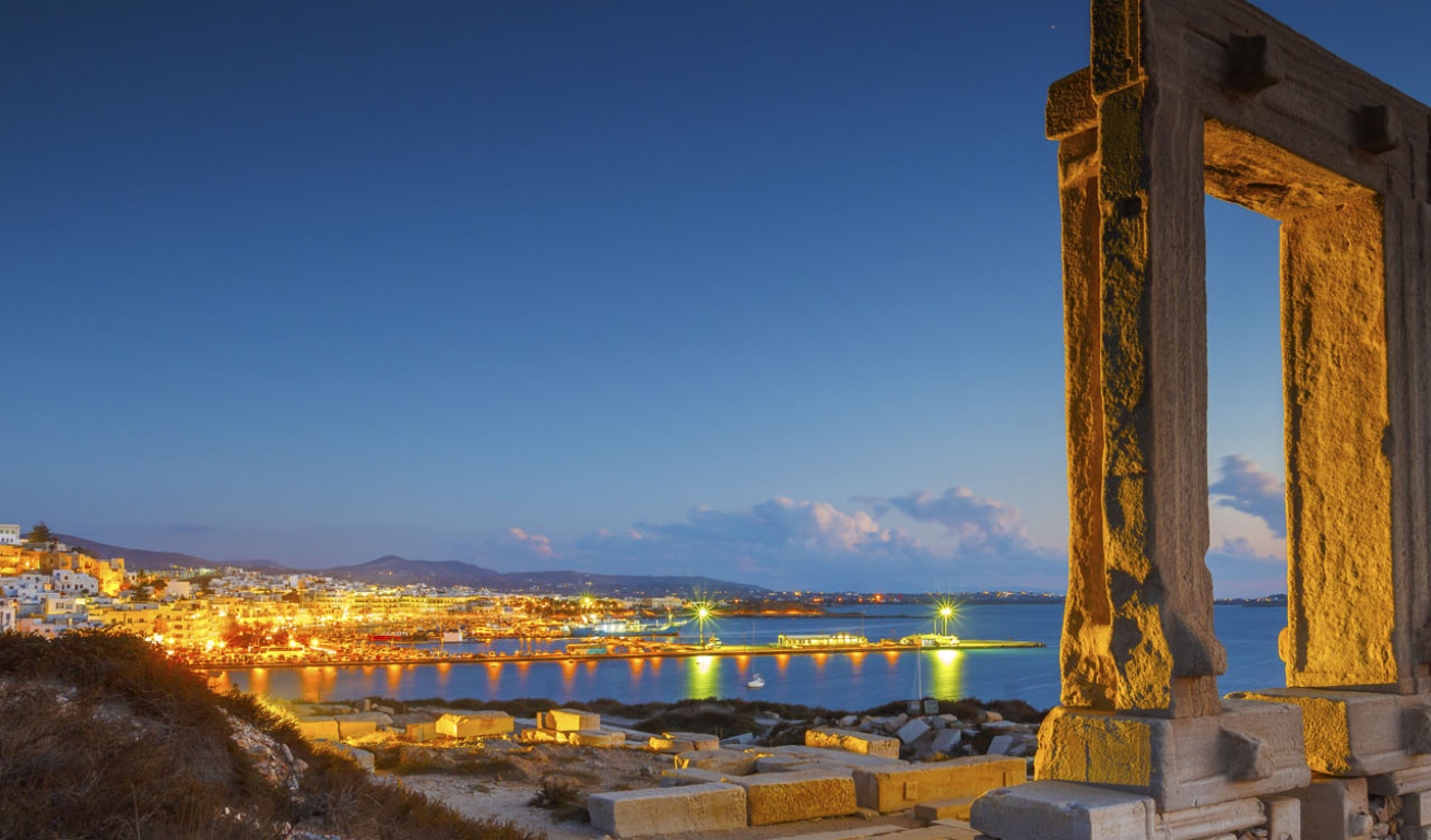Large ancient gate in the foreground and in the background a Cycladic settlement with a port at sunset. 