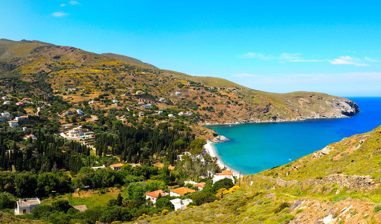 Cycladic landscape with low hills and α ravine with dense vegetation and small houses that ends up on a sandy beach.