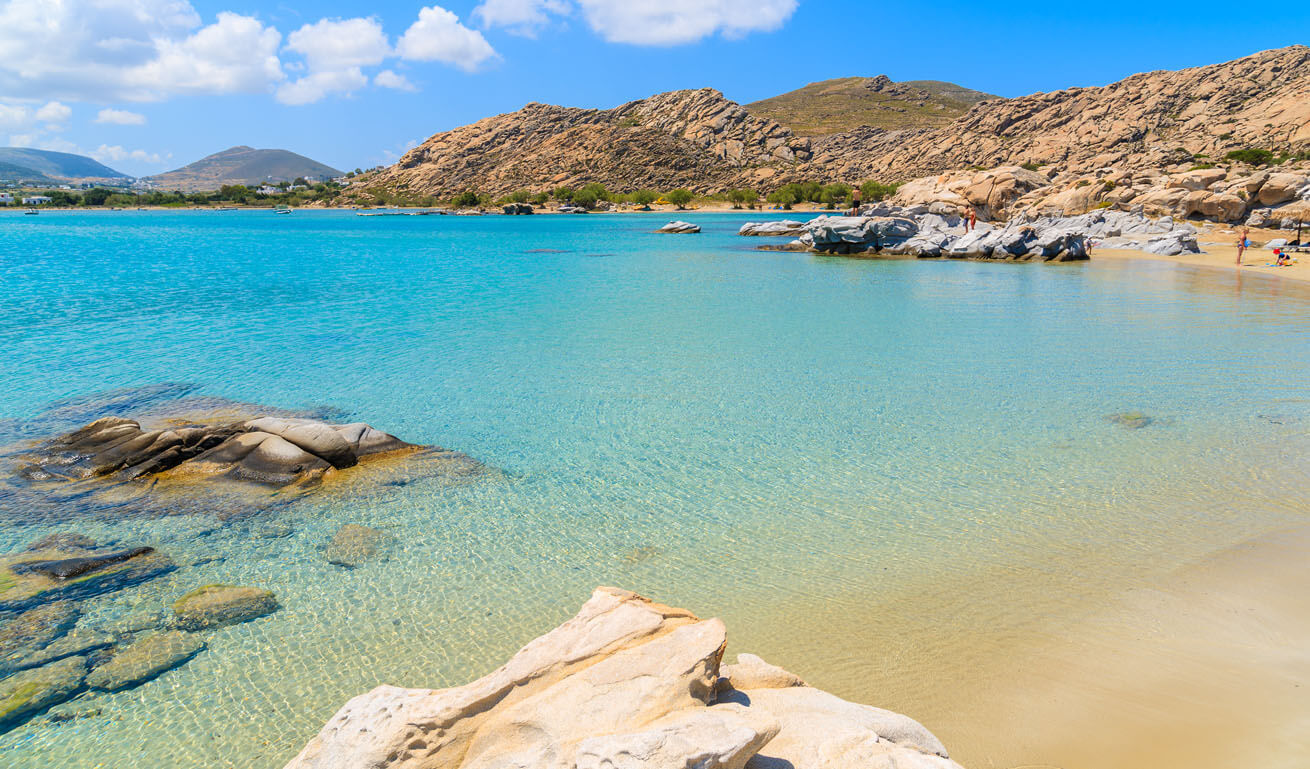 Yellow sandy beach with calm turquoise sea and a few small rocks. In the background other beaches and rocky hills. 
