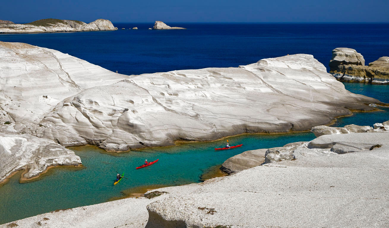 White rocks by the sea that form a deep bay. 3 canoes in a row proceed in the clear waters. 