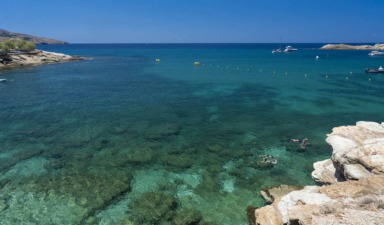 Shallow transparent sea where 4 people are swimming. Some rocks at the edge and in the background small yachts 