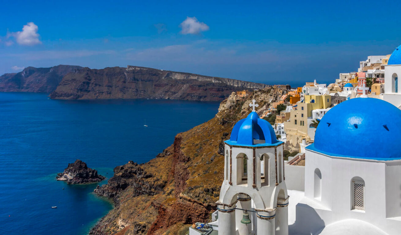 The domes of 2 churches and a settlement on a high rock above the sea and in the background 2 small islands nearby. 