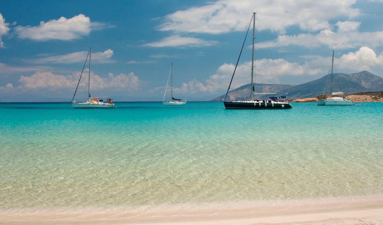 Beach with sand and turquoise waters and in the background sailing boats and land with high mountains. 