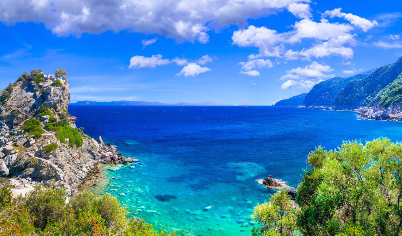 Ακτογραμμή με πράσινη βλάστηση και ένα μεγάλο βράχο στη θάλασσα. Στην κορυφή του μια μικρή εκκλησία. 