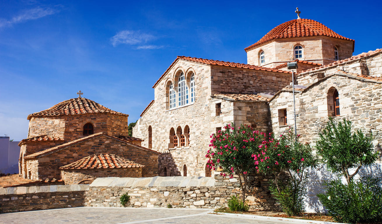 Two stone churches with a tiled dome: one large and one small and a few plants with pink flowers. 