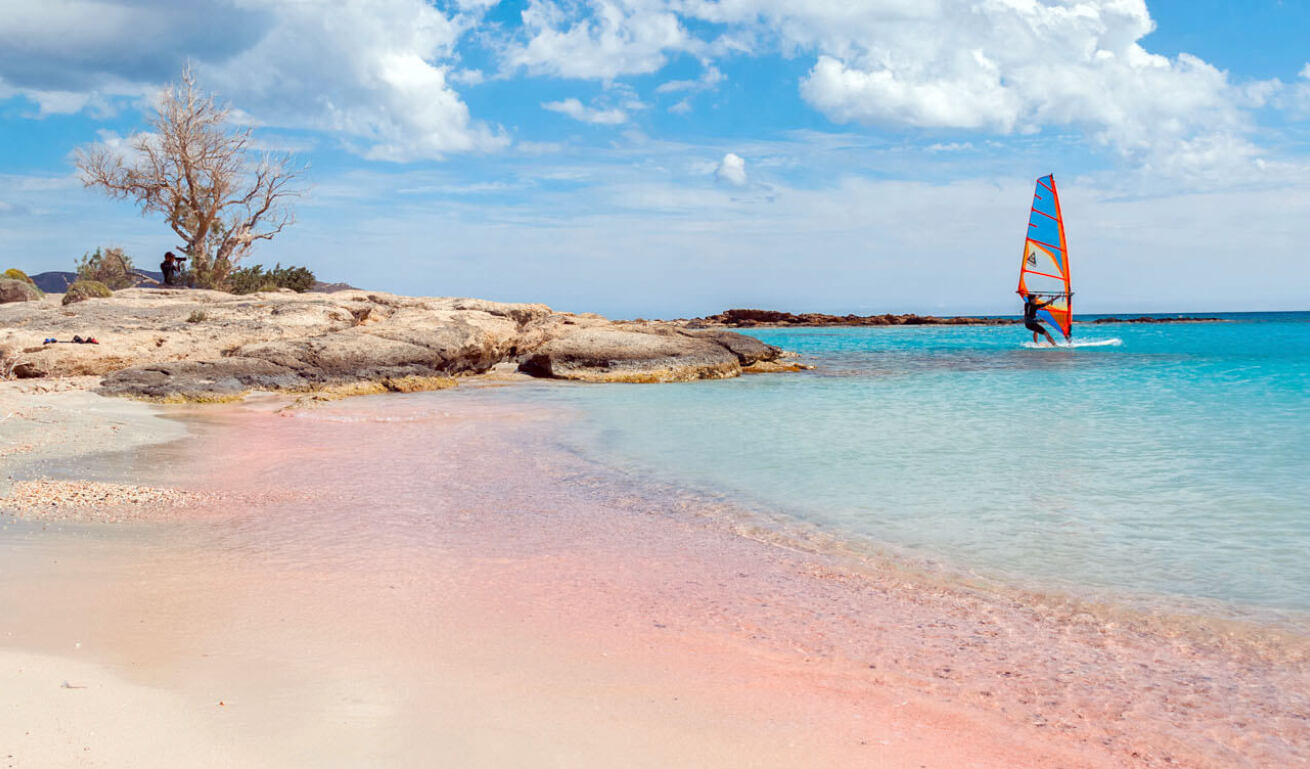 Beach with white and pink sand and low cliffs at the edge with little vegetation. A windsurf is sailing in the blue sea. 