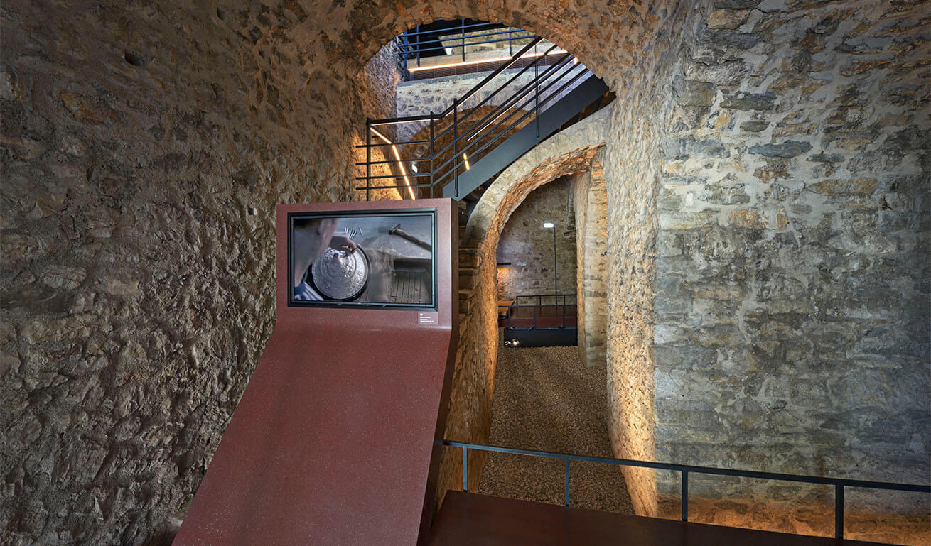 Interior of an old stone building with arches and stairs, transformed into a museum with a projection screen and special lighting.