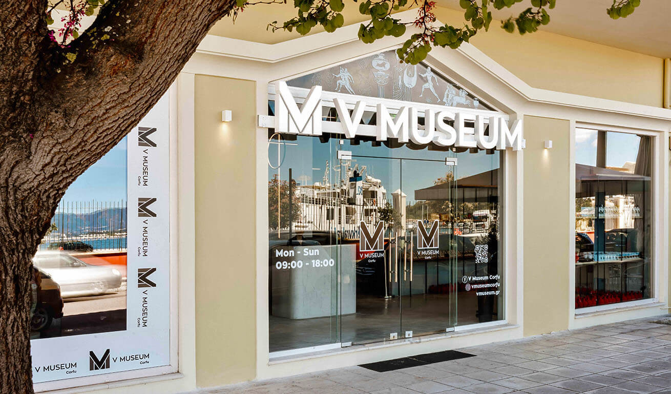 Γυάλινη πρόσοψη μουσείου με το όνομα του μουσείου, πλαισιωμένη από ανοιχτόχρωμο τοίχο. Αντανακλά χαμηλά κτίρια και καθαρό ουρανό.