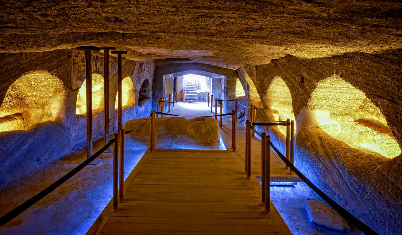 Underground wooden corridor inside a rock. At both sides illuminated recesses and in the background is the entrance of the space.