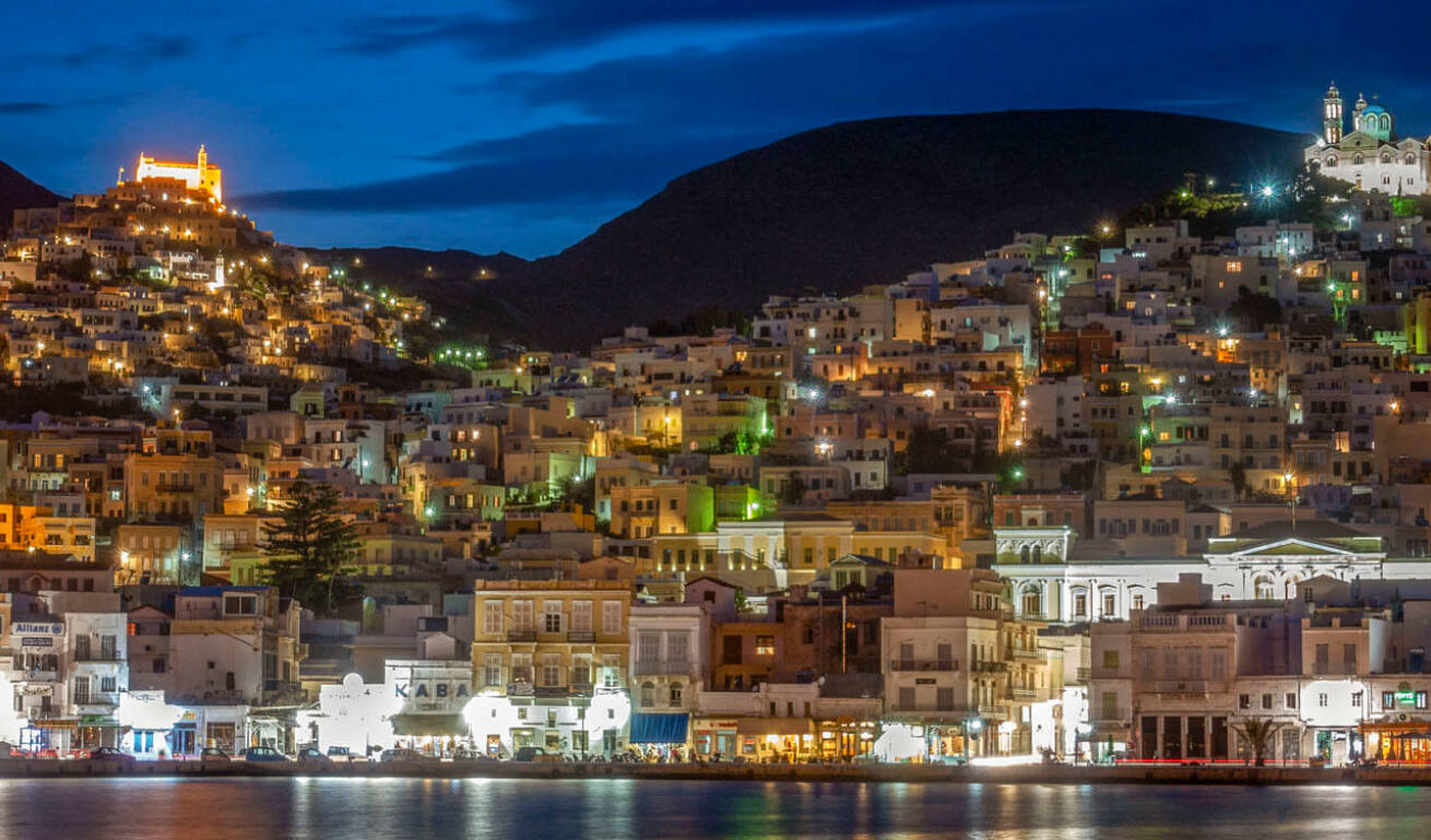 Night view of a large settlement from the sea with hills that have churches on the tops. 