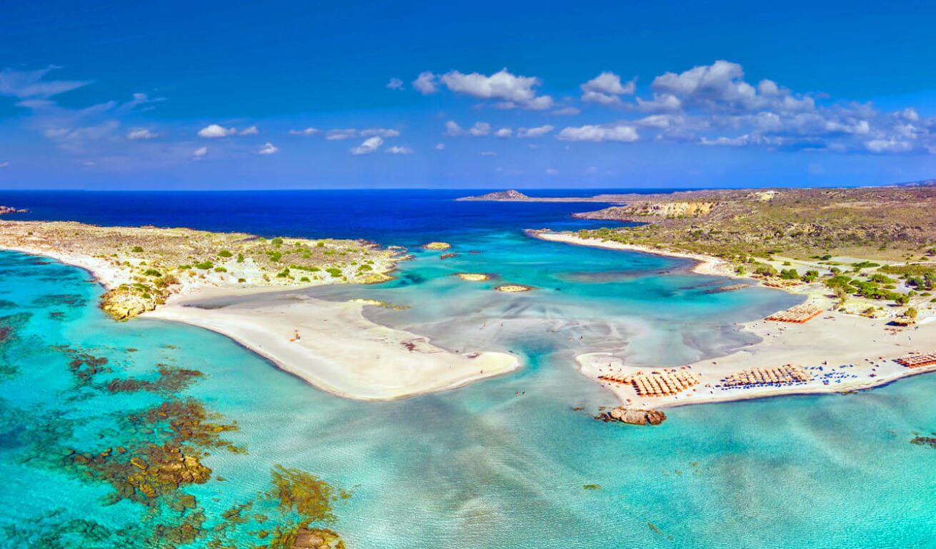 Aerial view of a coastal area with shallow waters, long sandy beach with some umbrellas and a small island near the beach.