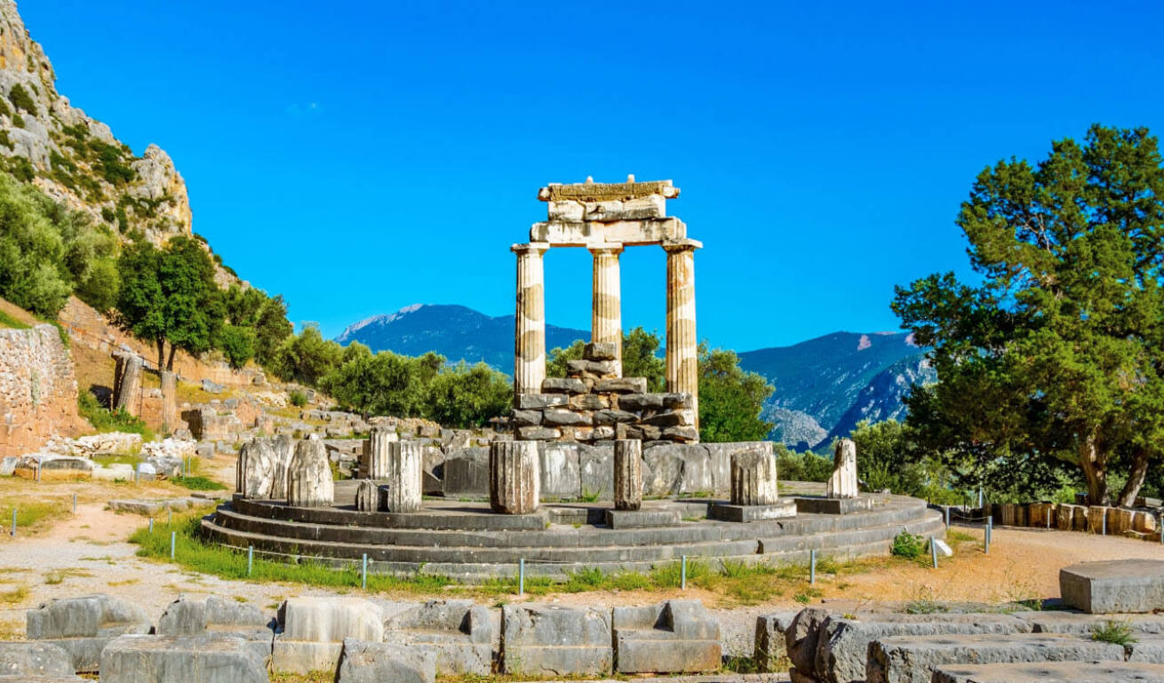 Archaeological site on a mountain slope with 3 upright columns and green trees around. 