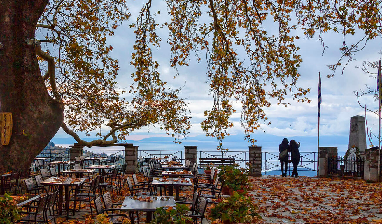 Άποψη  της κεντρικής πλατείας τιου χωριού, με δύο ανθρώπους να κοιτάζουν την καταπληκτική θέα και τραπέζια κάτω από τον πλάταννο