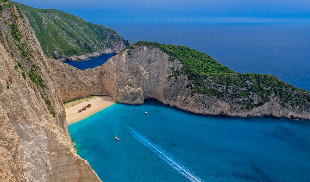 View from above of a sandy beach and a shipwreck. Around them are high rocks and in the background you can see the horizon.