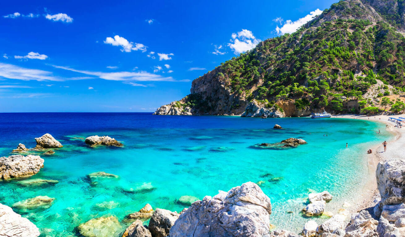 Παραλία με γαλαζοπράσινα νερά, βράχους και δέντρα, αλλά και λουόμενους που περιφέρονται