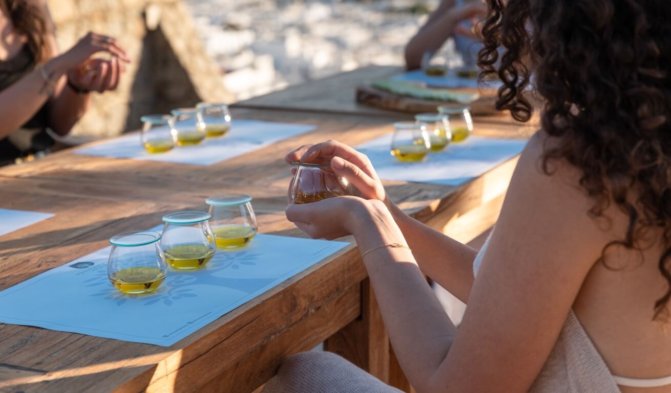 a woman sitting in front a table with 3 glasses of olive oil and holding a glass of olive oil, doing olive oil tasting