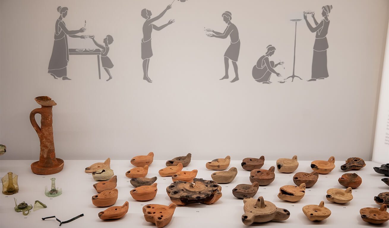 Έκθεση αρχαίων ελληνικών αγγείων και λύχνων, τοποθετημένων σε τραπέζι μπροστά από μια τοιχογραφία που απεικονίζει διάφορες μορφές να συμμετέχουν σε καθημερινές δραστηριότητες, σε μουσειακή έκθεση.