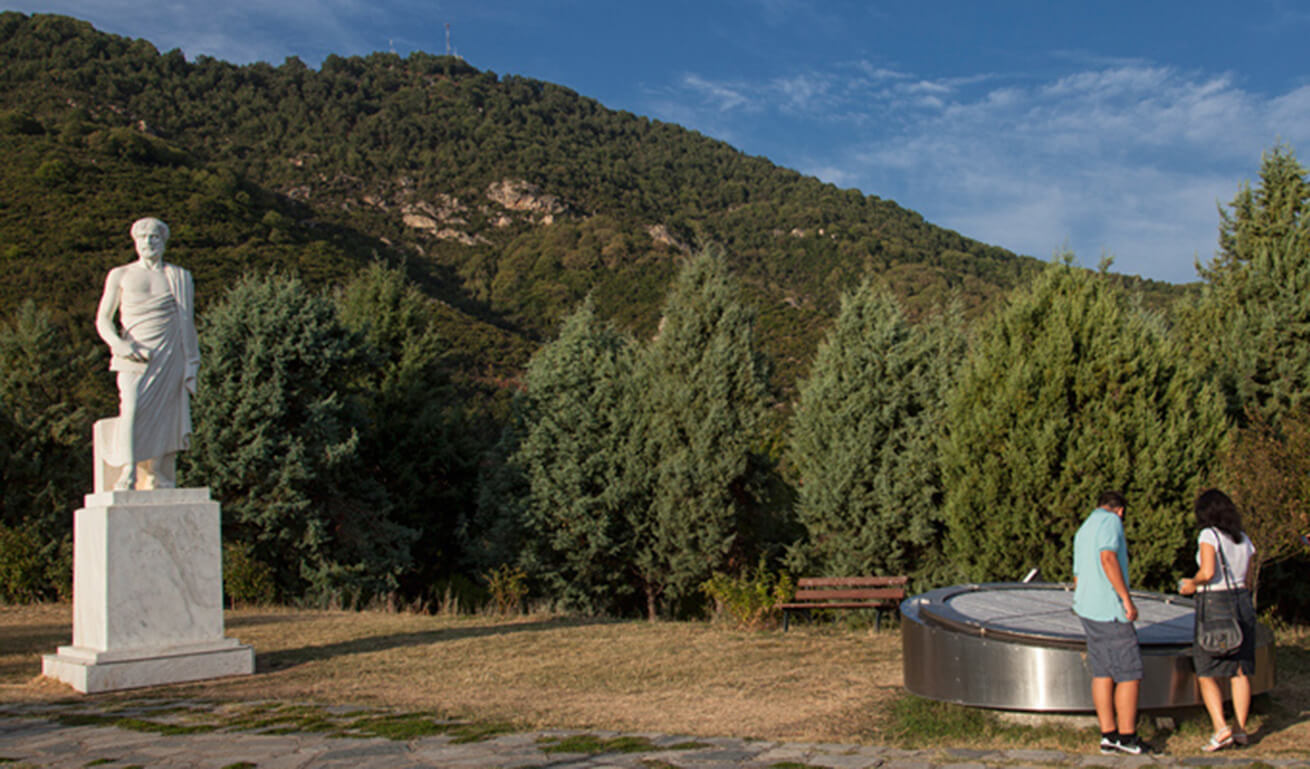 Two people standing beside a statue of Aristotle in a park, with dense trees and a hill in the background.

