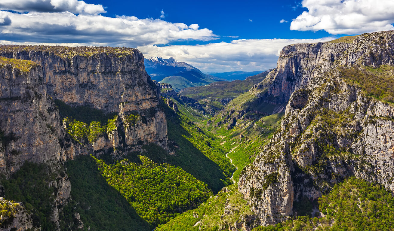A panoramic view of Vikos Gorge in Greece, featuring steep rocky cliffs and a lush green valley under a partly cloudy sky.