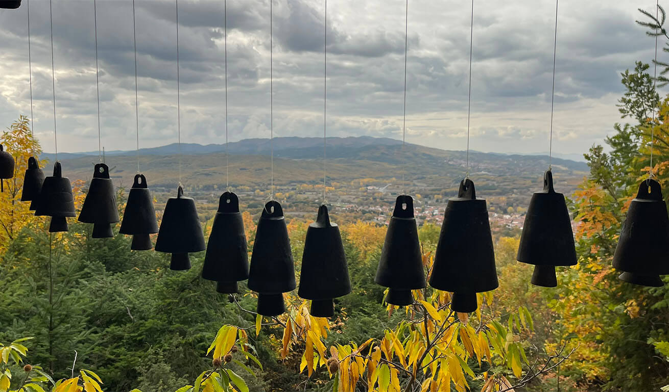 Row of dark bells hanging with strings against a scenic backdrop of mountains and a city, surrounded by autumn foliage.