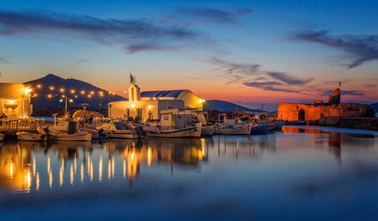 Peaceful harbor with boats and a small white church with lights at sunset.