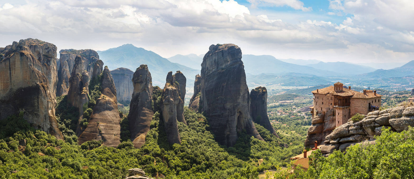 A panoramic view of high cliffs over a green valley. In the foreground there is a traditional stone monastery.