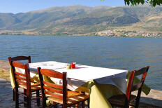 Τραπέζι με ξύλινες καρέκλες δίπλα σε λίμνη. Στο βάθος βουνά και παραλίμνιος οικισμός. Καλοκαίρι με λιακάδα.