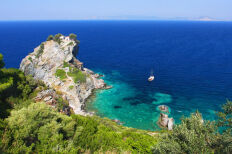 Ακτογραμμή με πράσινη βλάστηση και ένα μεγάλο βράχο στη θάλασσα. Στην κορυφή του μια μικρή εκκλησία. Στη θάλασσα ένα σκάφος.