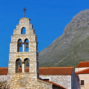 Tall stone-built belfry with 4 bells in front of tiled roofs of houses. In the background steep mountain slope.
