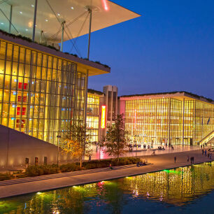 Modern glass building, lit from the inside with yellowish light, on the one side on the roof a large structure - a canopy supported by thin metal columns, people walking in the outer space and in front there is water from an artificial canal in which the building is reflected. Dusk.
