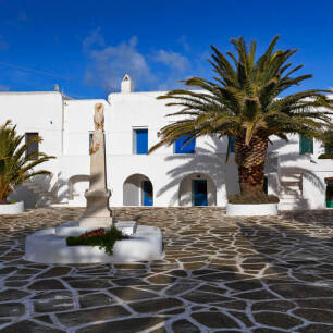 Πλακόστρωτη πλατεία με μια στήλη, φοίνικα και άσπρα σπίτια με καμάρες.