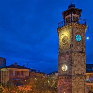 Tall stone clock in a square. In the background, a traditional stone house with a tiled roof. Night with lights.