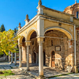 Byzantine stone church with dome and arches in the courtyard. Paved yard with small tree. 