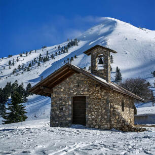 A small stone-built church with a snowy mountain behind in Kalavryta